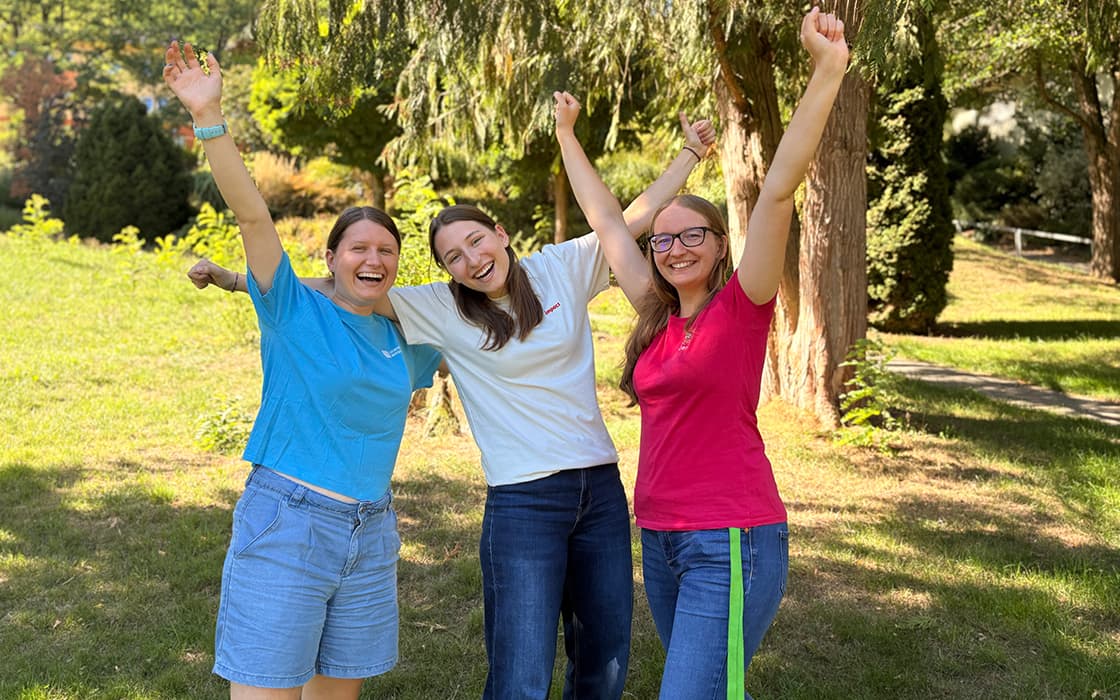 Drei lächelnde Frauen stehen draußen mit erhobenen Armen und feiern gemeinsam an einem sonnigen Tag auf einer Wiese.