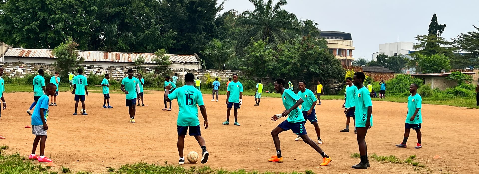 Eine Gruppe junger Männer in blauen Trikots spielt Fußball auf einem unbefestigten Platz, der von Bäumen und Gebäuden umgeben ist.