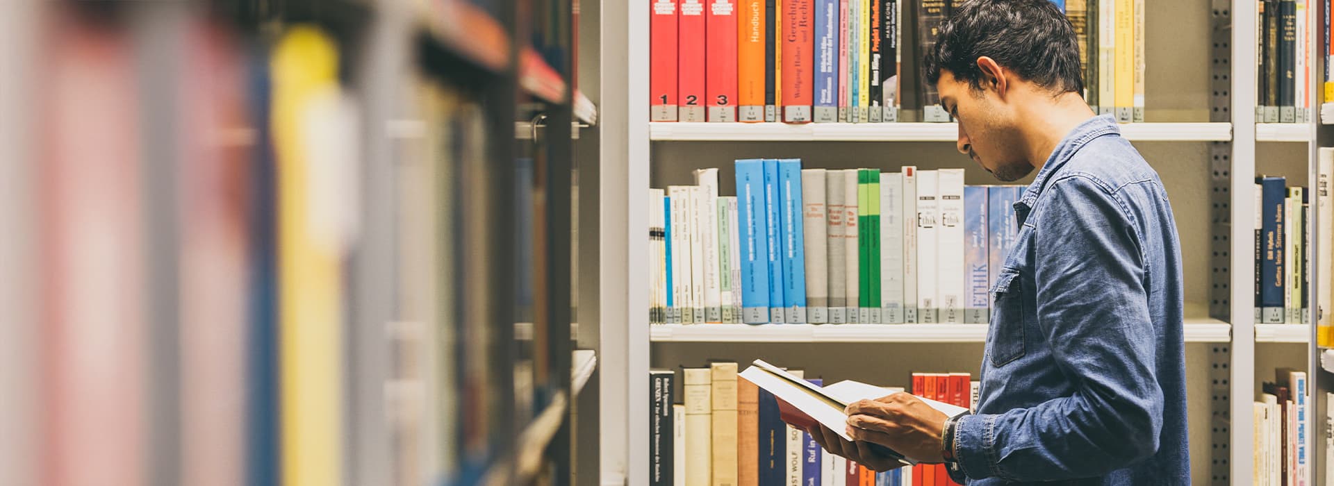 Ein Mann in einem Jeanshemd steht in einem Bibliotheksgang und liest ein Buch, hinter ihm stehen Regale mit bunten Büchern.