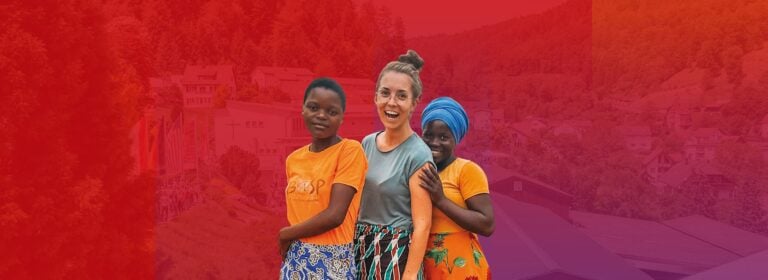 Drei lächelnde Frauen, zwei davon in orangefarbenen Oberteilen und Röcken, feiern das Pfingstmissionsfest vor einem farbenfrohen roten Hintergrund mit Farbverlauf.