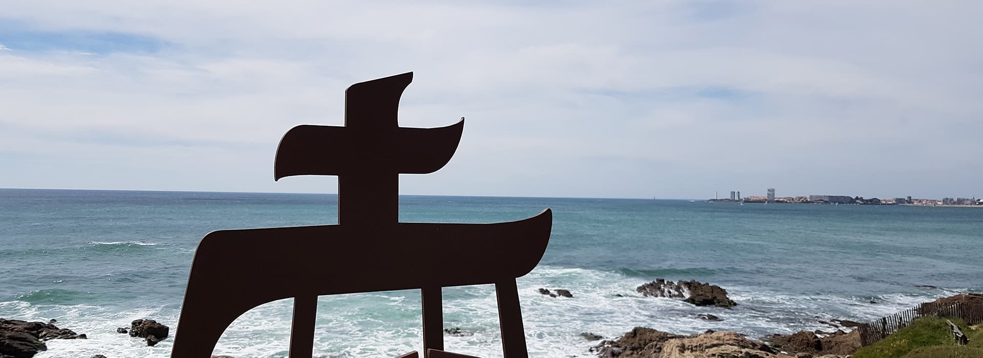 Metallskulptur eines stilisierten Bootes am Meer in Nizza, felsiges Ufer und entfernte Stadt unter einem teilweise bewölkten Himmel.