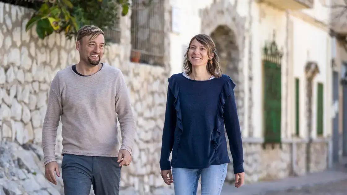 Daniel und Rosita Suchalla gehen draußen an einer Steinmauer in Spanien entlang, lächeln und genießen den sonnigen Tag.