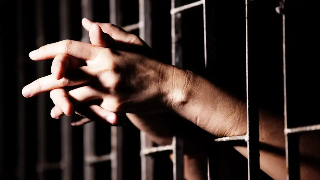Hinter metallenen Knaststäben verschränkte Hände in schummriger Beleuchtung, die an Gefangenschaft denken lassen.