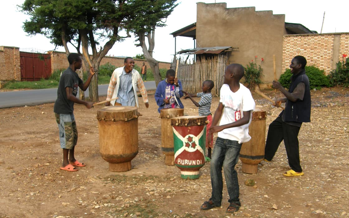 Sechs Jungen stehen im Freien und trommeln auf großen burundischen Trommeln in der Nähe eines Gebäudes und einer unbefestigten Straße bei Projekt Leben.