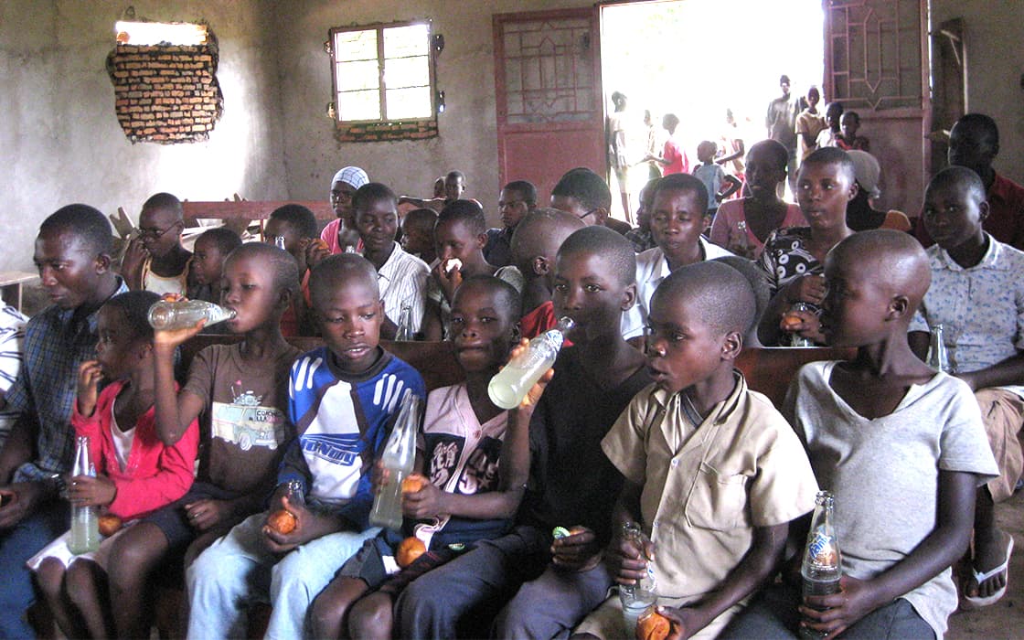 Eine Gruppe von Kindern in einem rustikalen Raum in Burundi, die Getränke und Snacks genießen, während die Erwachsenen von Projekt Leben hinter ihnen sitzen.