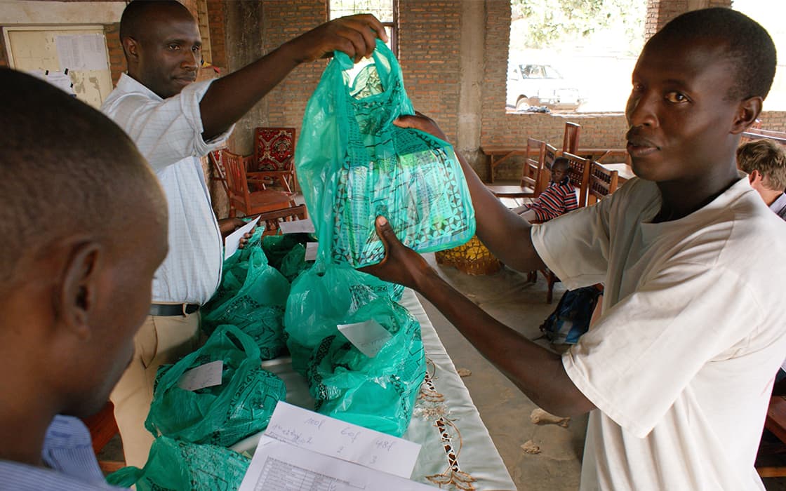 Ein Mann reicht einem anderen eine grüne Plastiktüte an einem Tisch mit Taschen und einem Dokument während des Projekts Leben in Burundi.