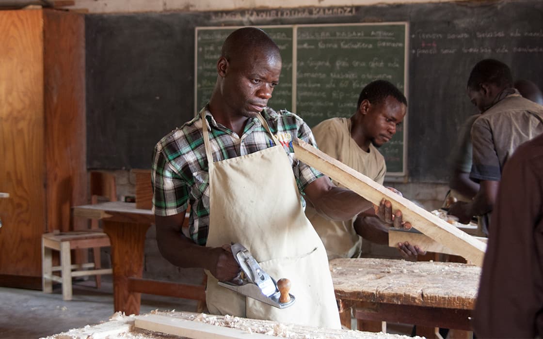 Ein Mann in einer Schürze bearbeitet im Chisomo-Ausbildungszentrum in Malawi Holz mit einem Handhobel, während andere in der Nähe arbeiten.