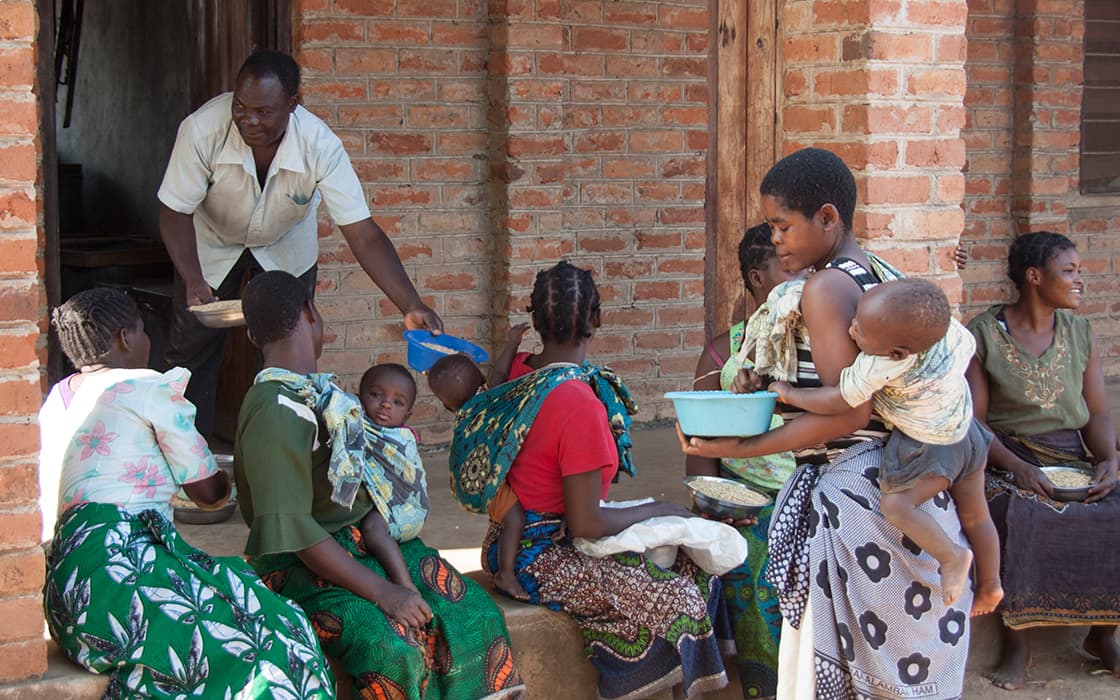 Frauen und Kinder sitzen vor einem Backsteingebäude in Malawi, während ein Mann im Chisomo-Ausbildungszentrum Essensschalen ausgibt.