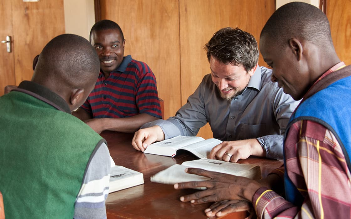 Vier Männer sitzen im Chisomo-Ausbildungszentrum um einen Tisch herum und lesen gemeinsam Bücher, lächelnd in einem warm beleuchteten Raum.