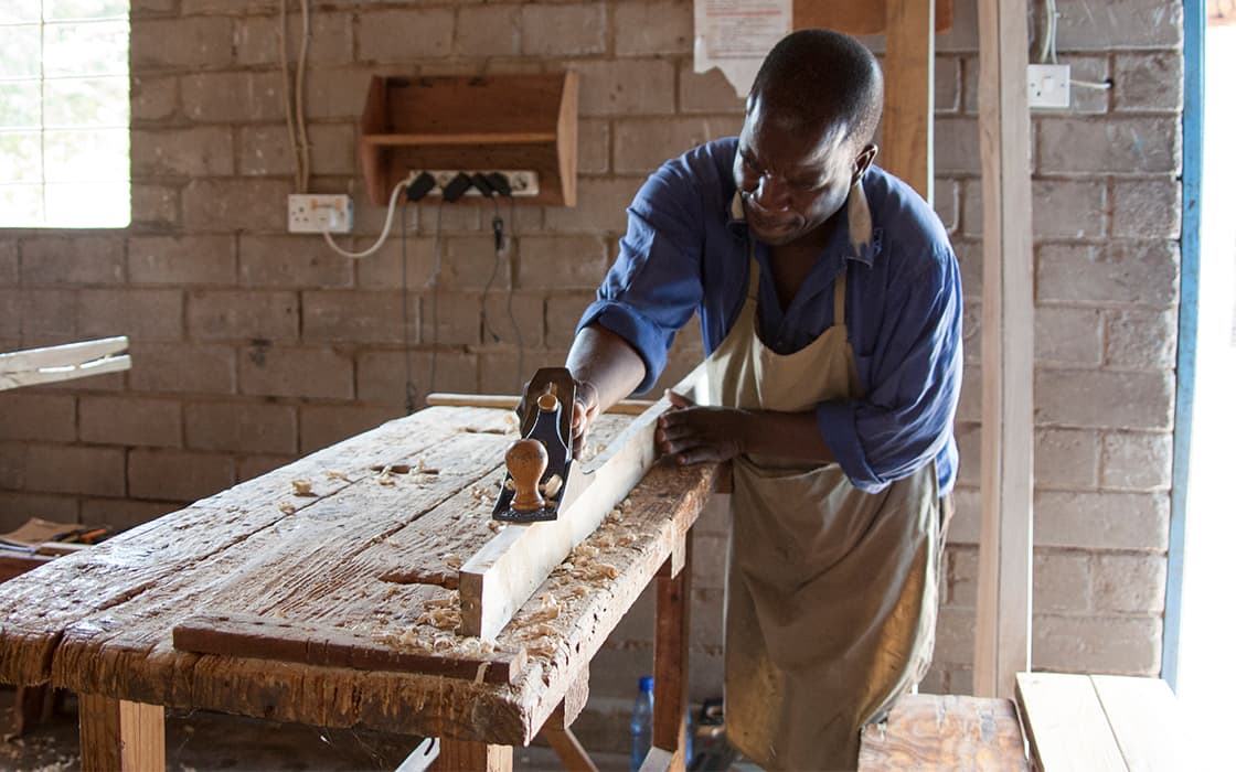 Ein Mann in einer Schürze bearbeitet Holz mit einer Handhobelmaschine im Chisomo-Ausbildungszentrum in Malawi, das von Ziegelmauern umgeben ist.