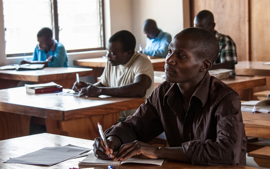 Mehrere Erwachsene sitzen im Chisomo-Ausbildungszentrum in Malawi an hölzernen Schreibtischen, schreiben auf Papier und konzentrieren sich auf ihre Arbeit.