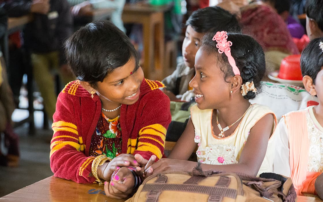 Zwei junge Mädchen im Kinderdorf Dinajpur in Bangladesch sitzen an einem Tisch, lächeln und unterhalten sich mit ihren Klassenkameraden.