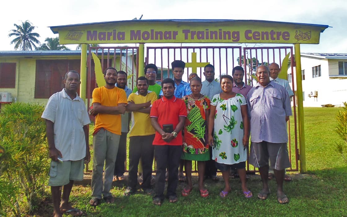 Eine Gruppe lächelt an einem sonnigen Tag vor dem gelben Tor des Maria Molnar Bible Training Centre in Papua-Neuguinea.