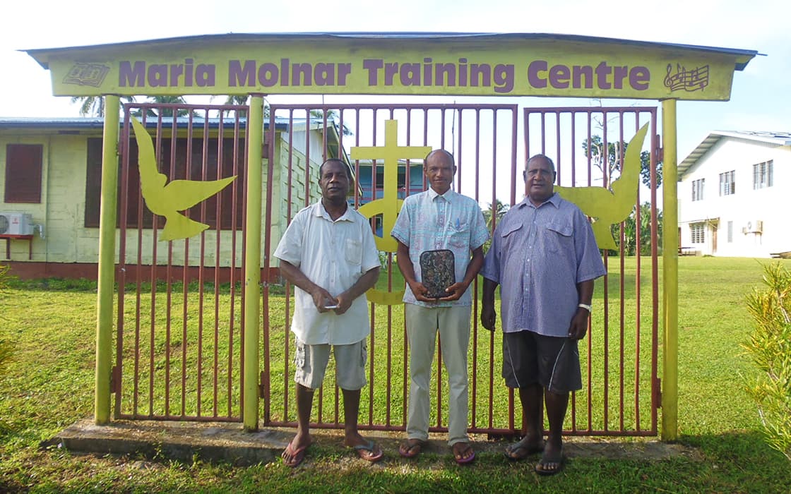 Drei Männer lächeln vor dem gelben Tor des Papua-Neuguinea Bible Training Centre auf einer Wiese in die Kamera.