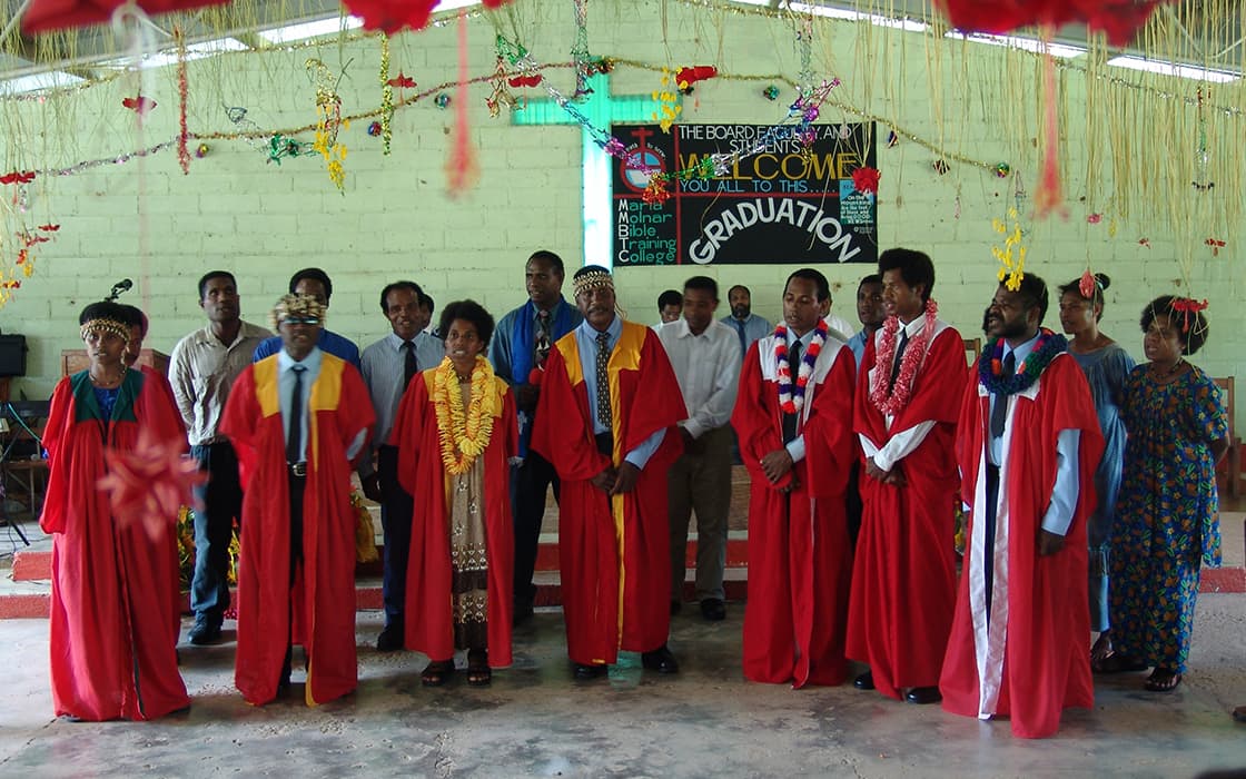 Eine Gruppe in roten Abschlusskleidern steht im Innenbereich des Bibelschulungszentrums, mit Dekorationen und einem Abschlussschild im Hintergrund.