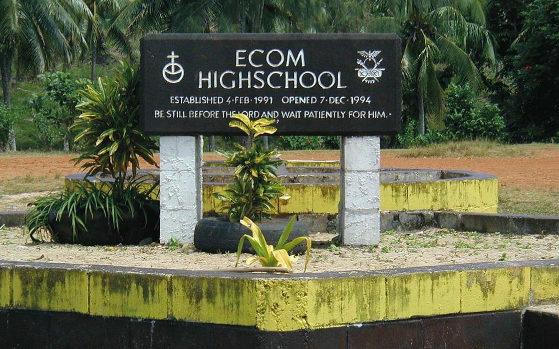 Ein schwarzes Schild für die ECOM High School und das Bibelausbildungszentrum, mit Daten, umgeben von Pflanzen in Papua-Neuguinea.
