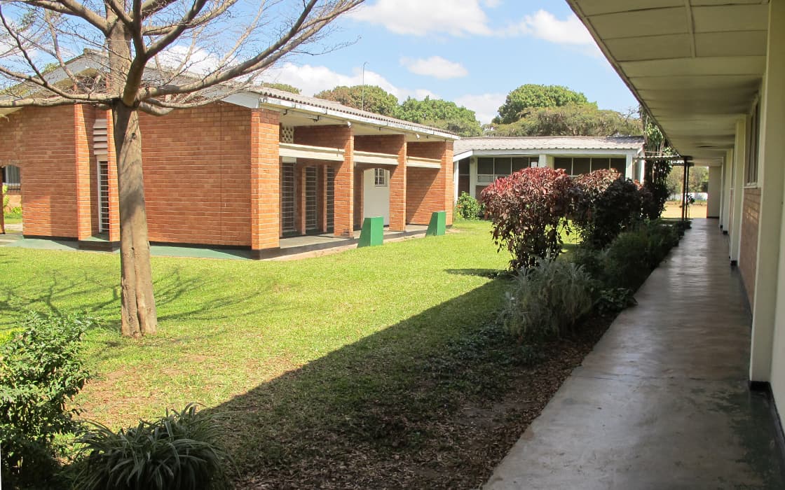 Ein Gehweg neben der Evangelischen Universität bietet einen Blick auf einen grasbewachsenen Hof mit roten Backsteingebäuden und Bäumen unter einem sonnigen Himmel.