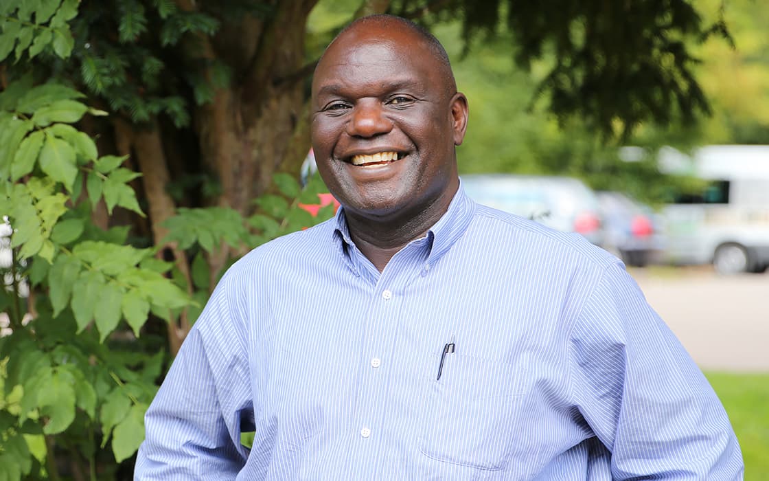 Ein lächelnder Mann in einem hellblauen Hemd steht im Freien an der Evangelischen Universität in Sambia, in der Nähe von Grünflächen und geparkten Autos.
