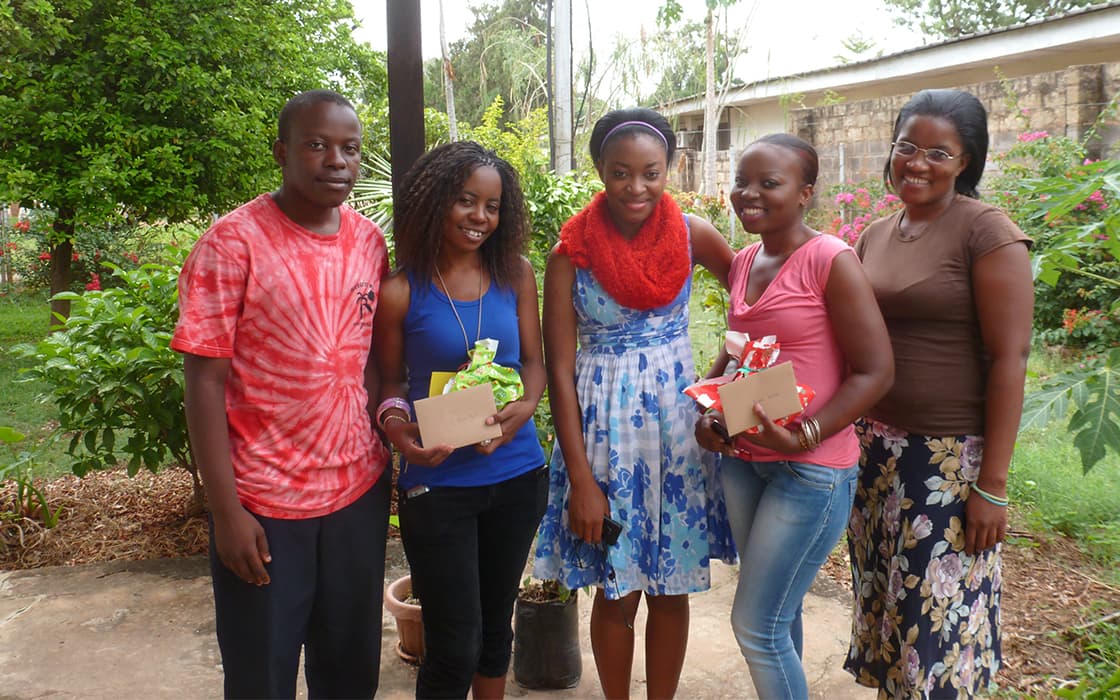 Fünf Personen stehen unter freiem Himmel an der Evangelischen Universität in Sambia, lächelnd mit Geschenken und Umschlägen inmitten von Grünpflanzen und einem Gebäude.