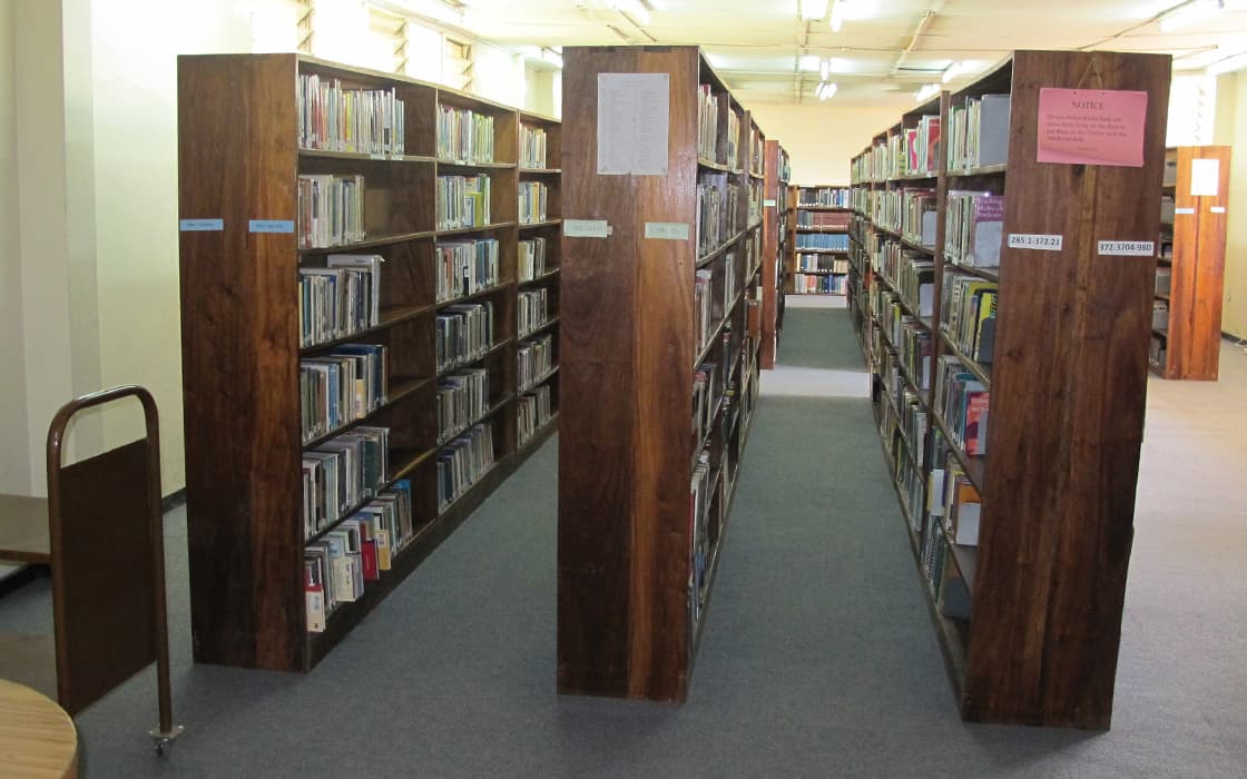 Holzregale voller Bücher säumen den mit Teppichboden ausgelegten und fluoreszierend beleuchteten Gang der Sambia-Bibliothek der Evangelischen Universität.