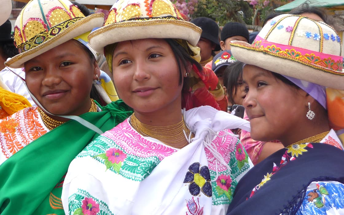 Drei Mädchen mit bunten Hüten lächeln während einer kulturellen Feier von Gemeindegründungen in Ecuador.