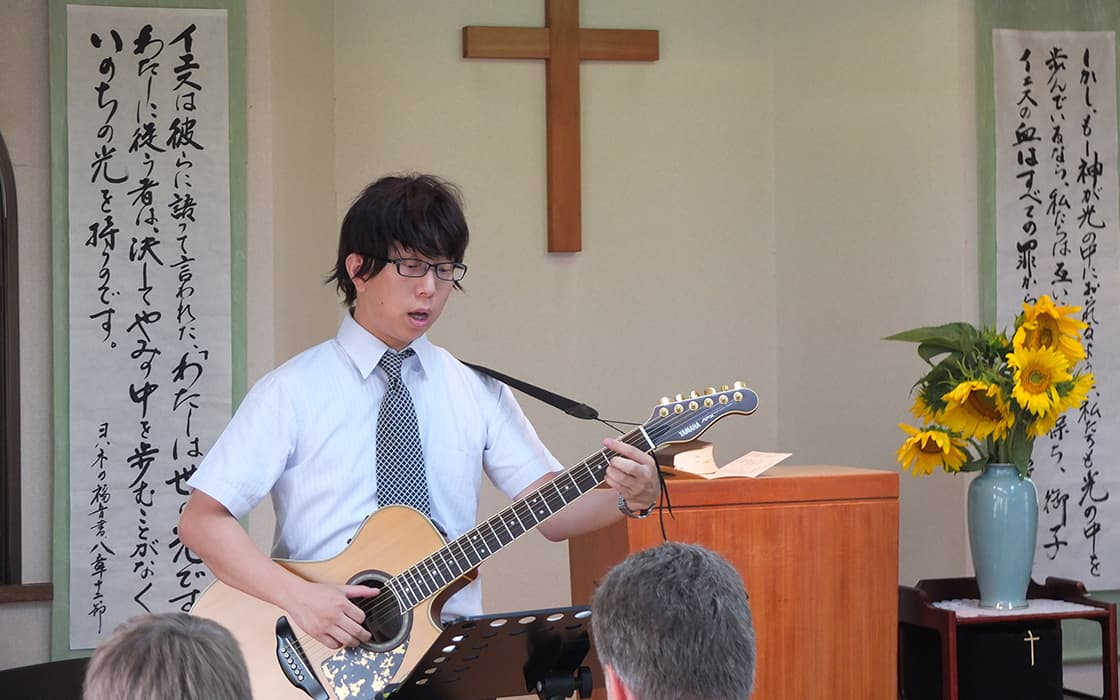 Ein Mann in Hemd und Krawatte spielt Gitarre und singt in einer Kirche, dahinter Gemeindegründungen Japan mit Kreuz und Sonnenblumen.