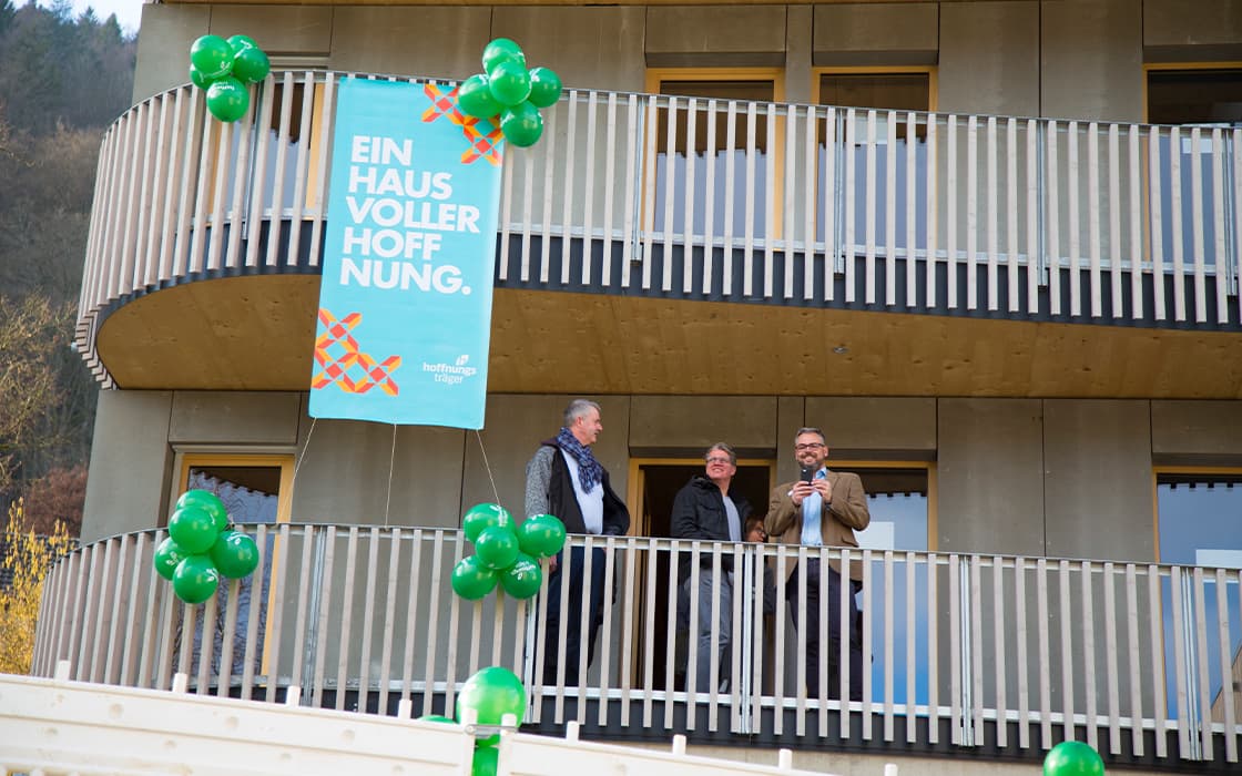 Drei Personen stehen auf einem Balkon in Bad Liebenzell mit grünen Luftballons und einem Hoffnungshaus-Banner mit der Aufschrift "Ein Haus voller Hoffnung".