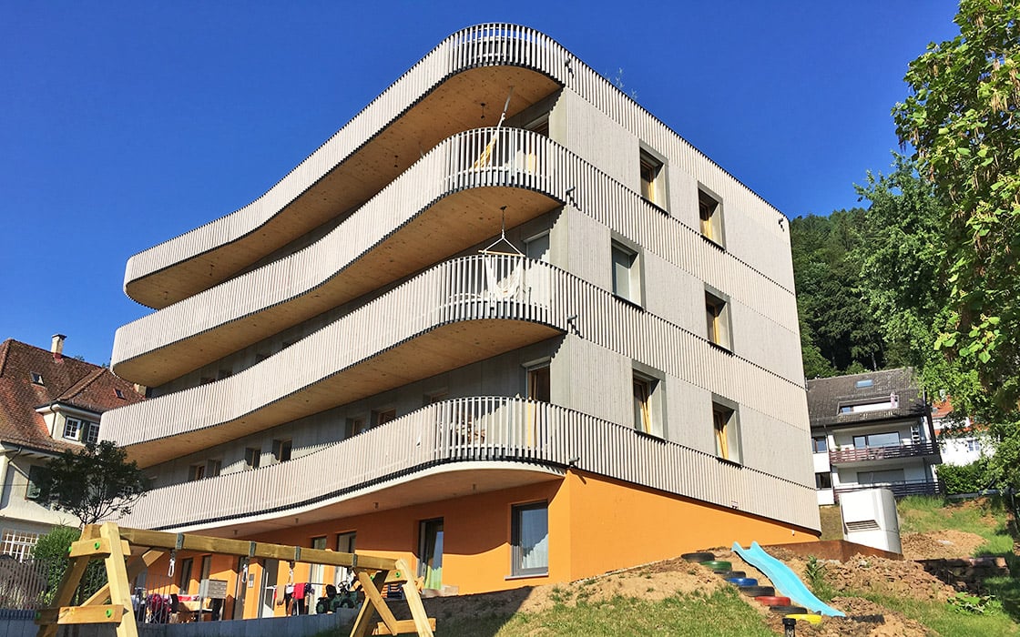 Modernes Gebäude in Bad Liebenzell mit geschwungenen Holzbalkonen, Orangenebene und einem Spielplatz auf einem Grashang.