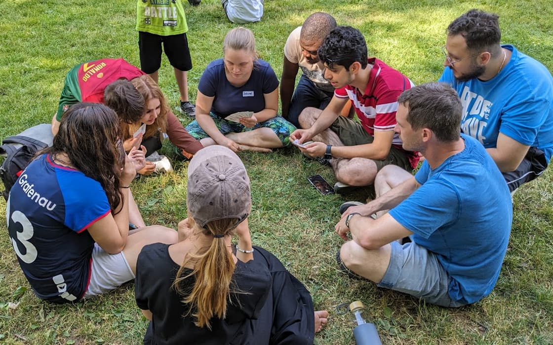 Eine Gruppe diskutiert über Integration, sitzt im Kreis auf einer Wiese im Freien und untersucht etwas in ihren Händen.