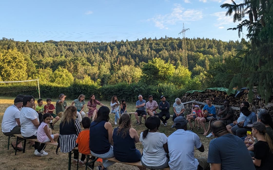 Eine Gruppe von Menschen sitzt in einem großen Kreis im Freien, umgeben von Bäumen, und genießt den Sonnenschein während eines Integrationseinsatzes.