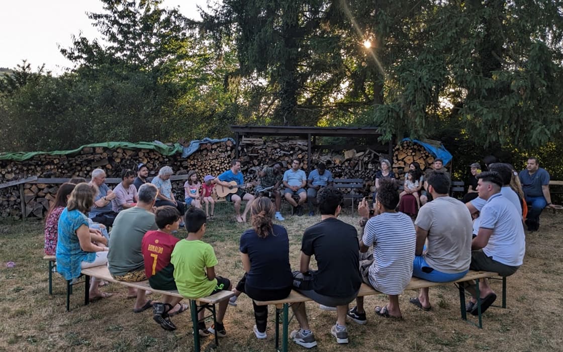 Eine Gruppe genießt ein sonniges Beisammensein in Deutschland, sitzt im Kreis am Feuerholz, einige spielen Gitarre und fördern so die Integration.