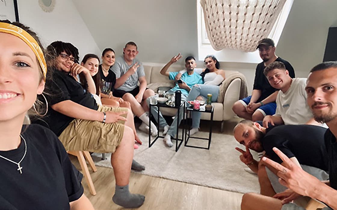 Eine Gruppe von elf lächelnden Freunden, die durch ihre Mission der Integration vereint sind, posieren für ein Selfie in einem gemütlichen Wohnzimmer.