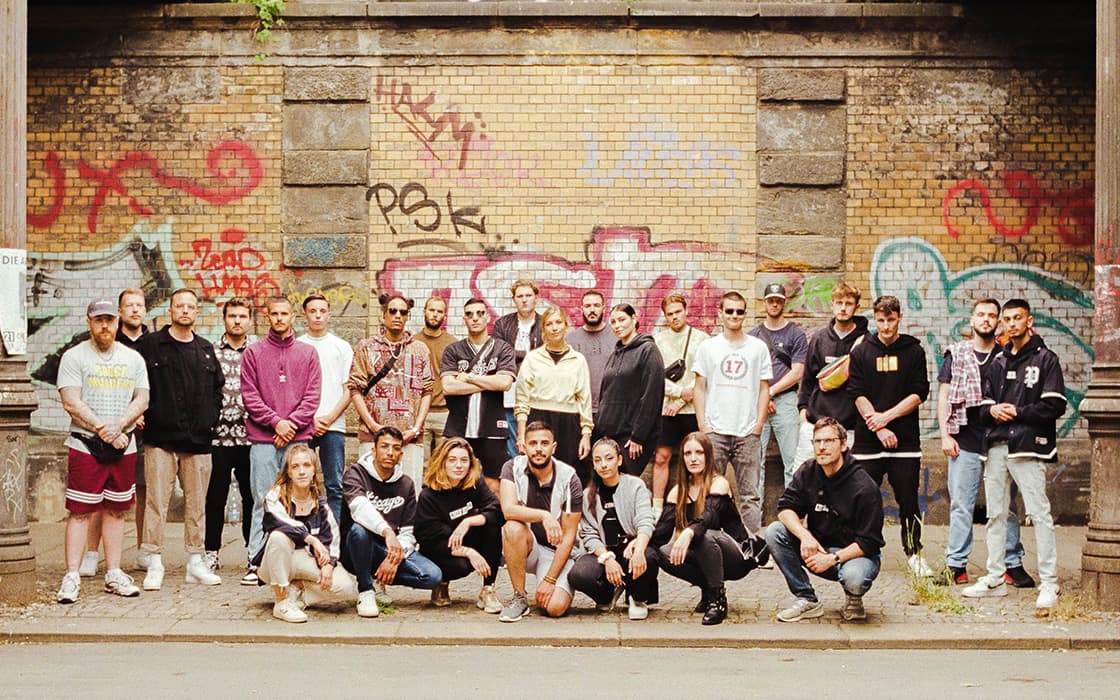 Vor einer mit Graffiti beschmierten Backsteinmauer unter einer Überführung posiert eine Gruppe von Menschen, die ihre Integration demonstrieren.