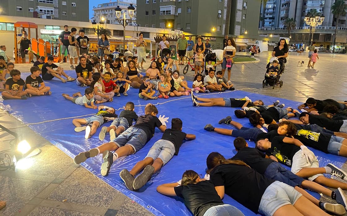 Kinder und Erwachsene versammeln sich zu einer Gruppenaktivität in Spanien, einige sitzen, einige liegen auf einer blauen Plane auf dem Platz.