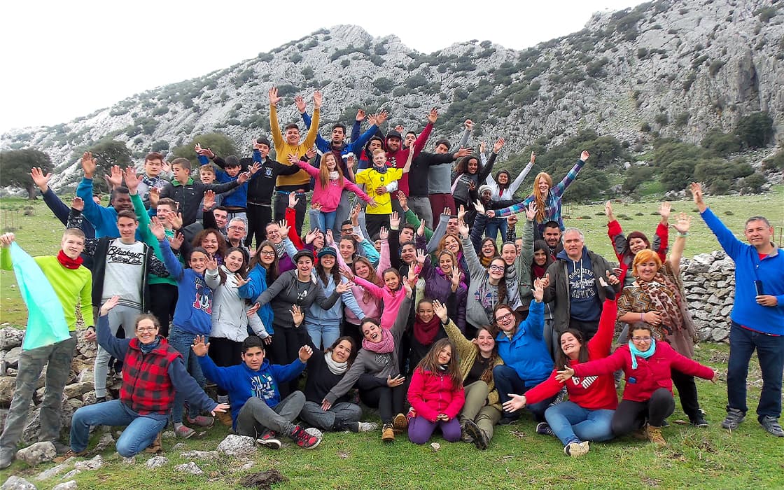 Eine große Gruppe posiert im Freien in Spanien, lächelnd und mit erhobenen Armen, mit Hügeln und Felsen im Hintergrund.