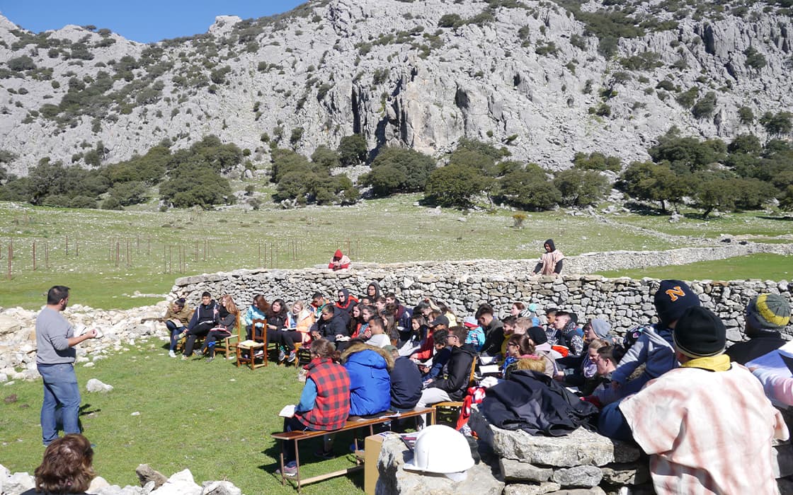 Eine Gruppe hört einem Redner im Freien an der Costa del Sol zu, mit felsigen Hügeln und Steinmauern im Hintergrund.