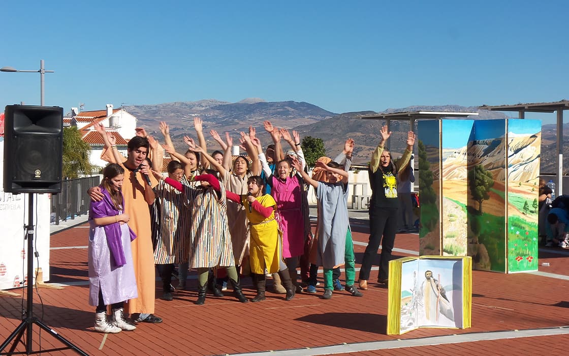 Eine Gruppe in farbenfrohen Kostümen tritt mit erhobenen Armen im Freien in Spanien auf, mit gemalten Kulissen und Bergen im Hintergrund.