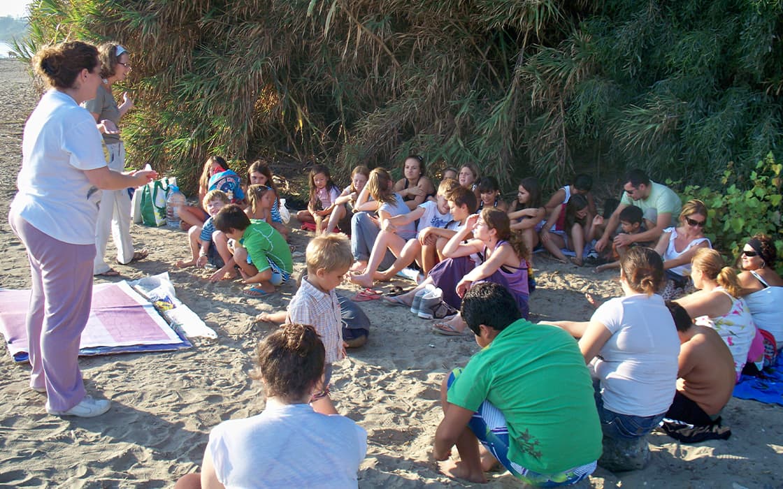 Eine Gruppe genießt die Gemeindearbeit an einem Sandstrand an der Costa del Sol und hört zwei Frauen in der Nähe von hohen Grünpflanzen zu.