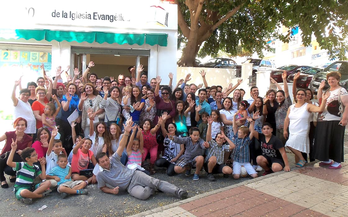 Eine große Gruppe von Menschen, darunter Kinder und Erwachsene, lächelt und winkt vor einer Kirche an der Costa del Sol.