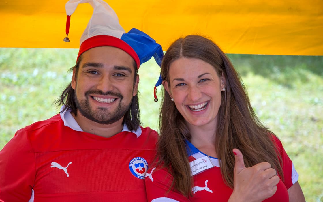 Zwei Menschen in roten Hemden - einer mit Narrenkappe - zeigen unter einem gelben Baldachin bei Mission Mobilizing Chile lächelnd die Daumen nach oben.