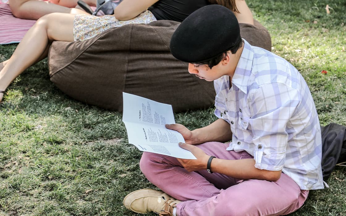 Eine Person mit schwarzer Baskenmütze sitzt in Chile auf einer Wiese und liest ein Buch; andere entspannen sich in der Nähe auf einer Decke und einem Sitzsack.