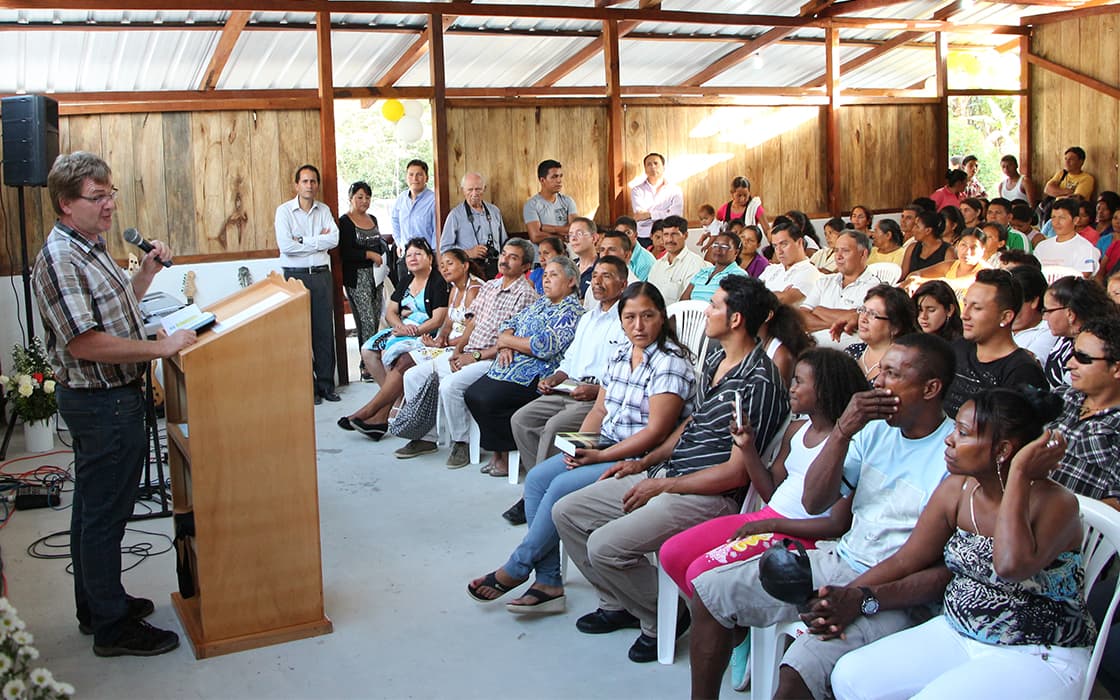 Ein Mann spricht an einem Podium über Mitarbeiterschulungen zu einem sitzenden Publikum in einem hellen, hölzernen Gemeindesaal.