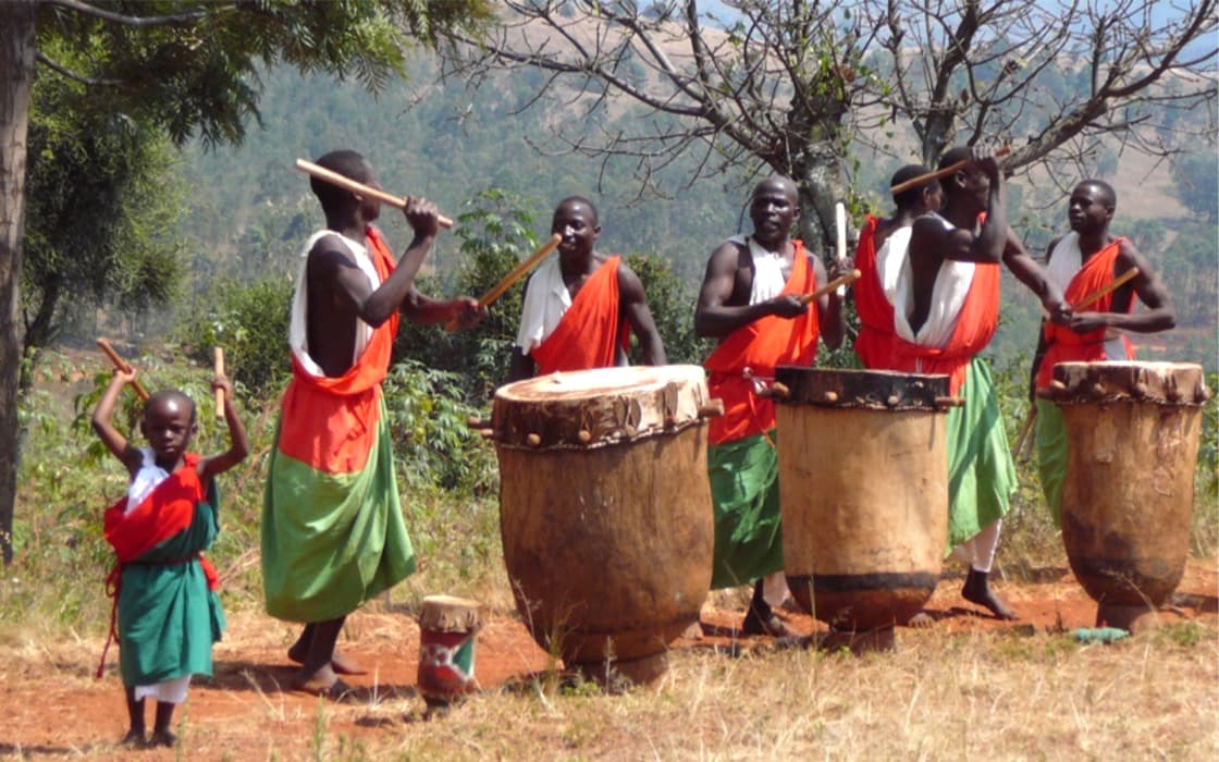 Sechs Personen, darunter ein Kind, in Rot, Weiß und Grün spielen im Freien in Burundi traditionelle Trommeln.