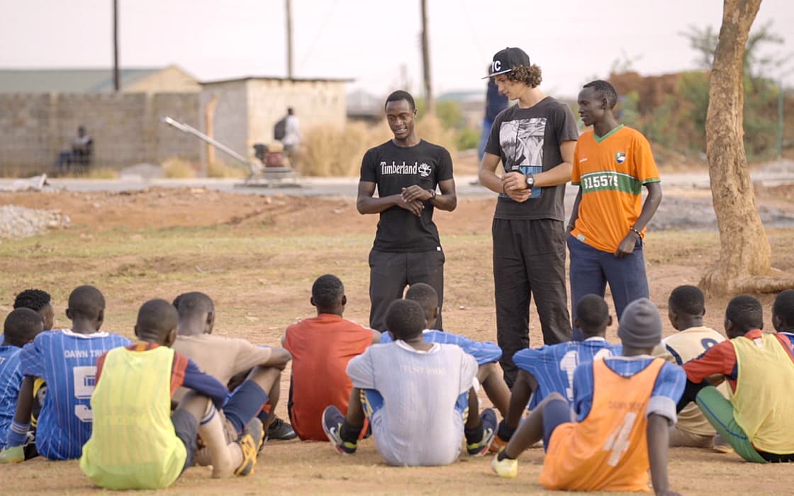 Drei Männer stehen und sprechen mit einer sitzenden Gruppe von Jungen in Sportkleidung auf einem unbefestigten Feld in Mushili, Sambia.