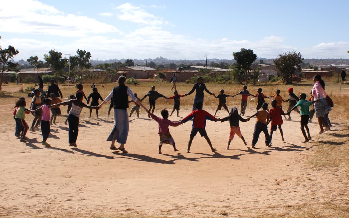 Eine Gruppe in Sambia genießt einen sonnigen Tag, hält sich an den Händen und spielt in einem großen Kreis mit Hilfe zum Leben.