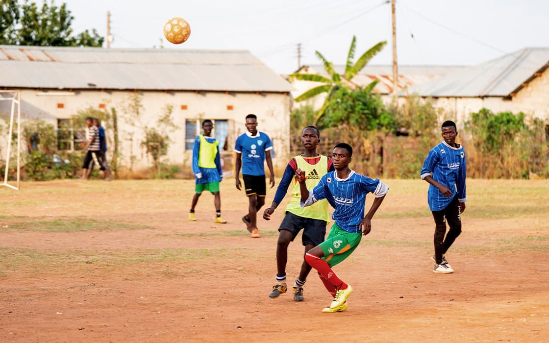 Jungen spielen Fußball auf einem staubigen Mushili-Feld in Sambia; ein Spieler, der von Hilfe zum Leben unterstützt wird, beobachtet den Ball in der Luft.