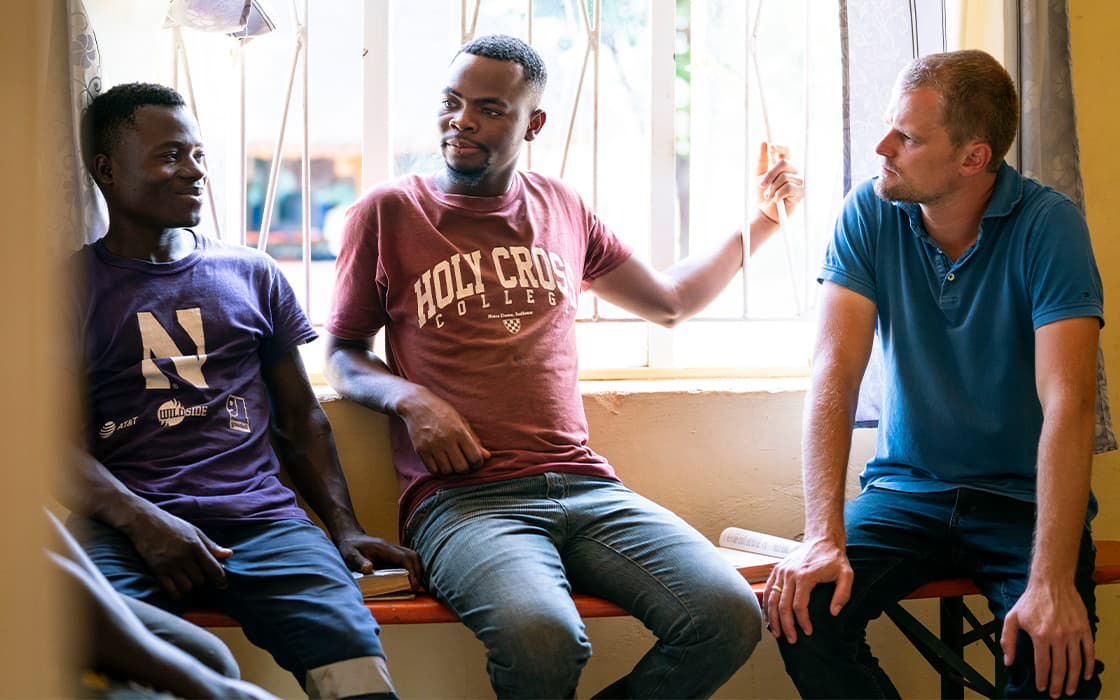 Drei Männer sitzen drinnen an einem Fenster in Sambia und unterhalten sich, während das Sonnenlicht den Raum füllt.