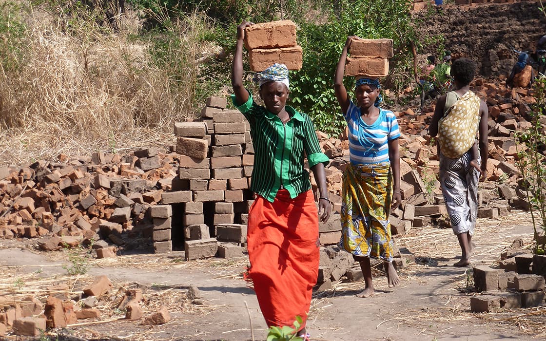 Zwei Frauen in Nabwalya, Sambia, tragen Ziegelsteine auf dem Kopf und suchen Hilfe inmitten von Ziegelstapeln und trockenem Gras.