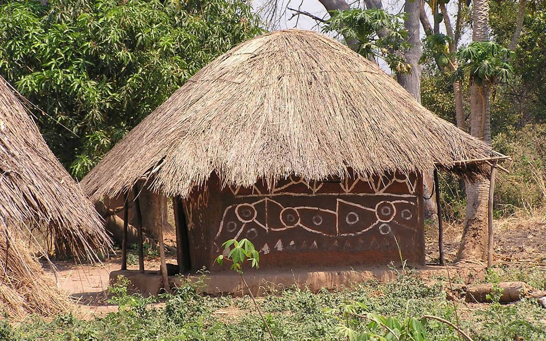 Eine runde Hütte mit Strohdach und geometrischen Mustern, typisch für Nabwalya, Sambia, steht inmitten von Bäumen und trockenem Gras.