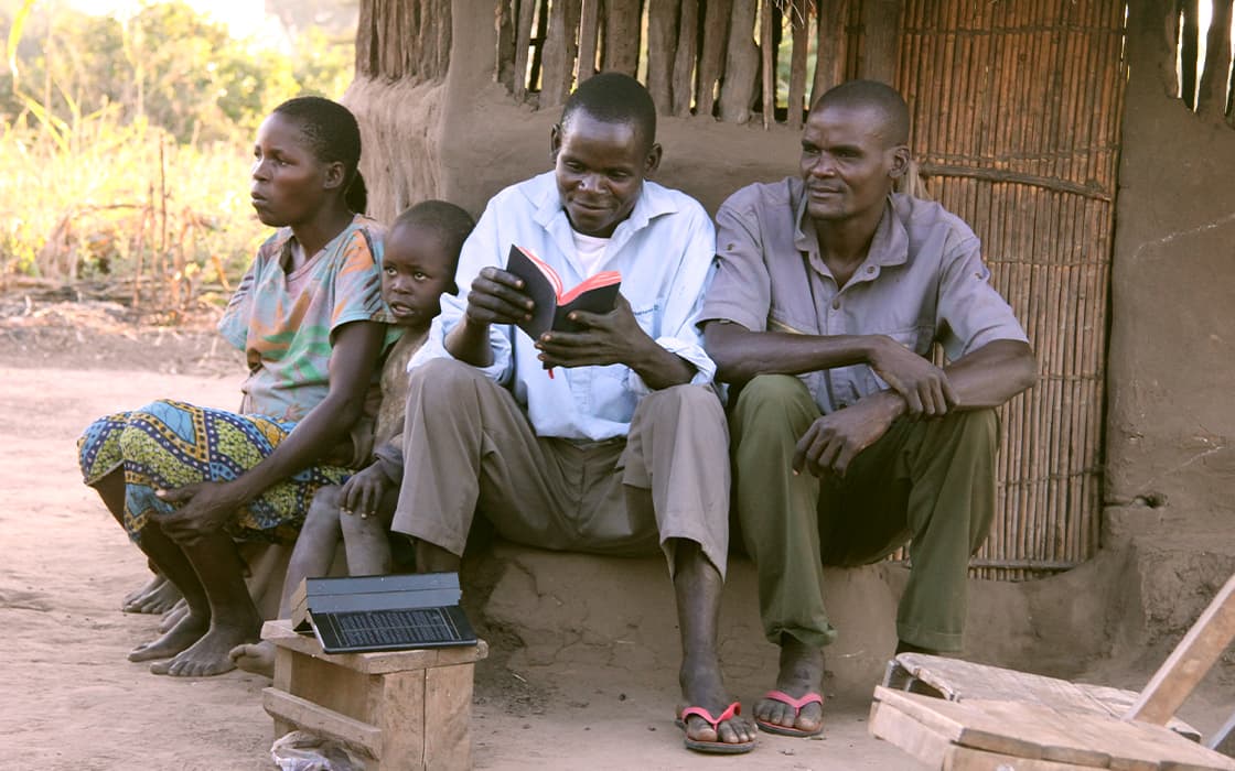 Vier Personen sitzen vor einem rustikalen Gebäude in Sambia; ein Mann liest, während die anderen zuschauen, mit einem Tablet auf einer Bank.